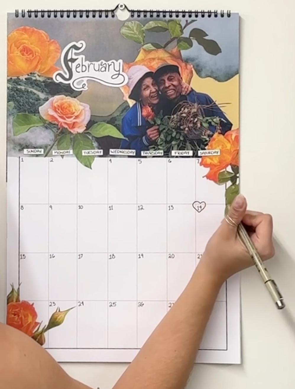 A spiral-bound February desk calendar with a floral rose design and a photo of a couple near the top, shown on a white wall with a hand holding a pen writing on the calendar grid.